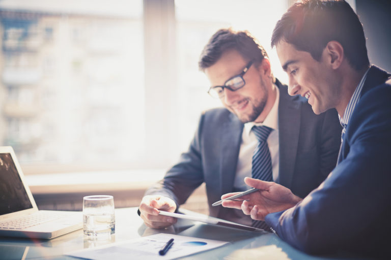 Two men in suits sit at a table while reviewing content together on a tablet.