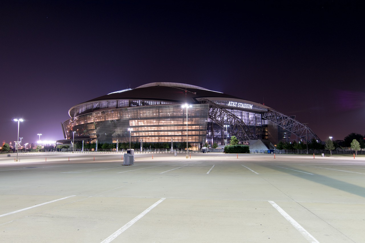 Exterior shot from an empty parking lot of a fully-lit AT&T Stadium at night.